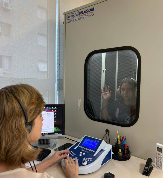Simone Ebel realizando avaliação auditiva com criança dentro da cabine audiométrica
